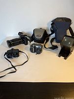 Overview of all items on tabletop: digital camera, film camera with lens, flash unit, binoculars, two camera bags