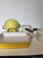 Side view showing yellow colander, rectangular baking dish and two white saucepans with wooden handles, all vintage enamelware with visible wear.