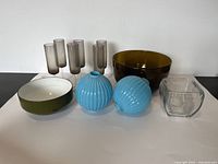 Full lot showing all items on a white sheet against white wall including green Copco bowl, six foggy grey glasses, amber bowl, clear square bowl, and two blue orbs.