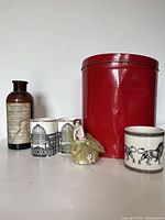 Photo showing entire lot with porcelain figurine, two glass cups, vintage bottle, and large red tin