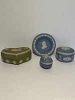 Front view of four Wedgwood Jasperware items: green heart-shaped box with white relief, blue round box, matching blue plate, and small egg-shaped blue box.
