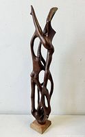 Front view of tall, slender abstract wood sculpture depicting intertwined nudes, mounted on a light wood base, with dark polished finish.