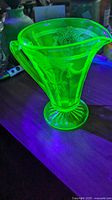Green Vaseline glass cream jug glowing under UV light showing fluorescence and etched Art Deco design.