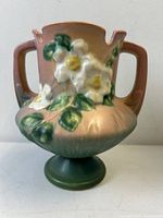 Front angle of Roseville art pottery vase showing white rose floral relief design, two handles, pedestal base with pink and green glazing.