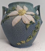 Front view of Roseville Clematis pottery vase showcasing white clematis flower and green leaves on blue-green textured background with dual handles.