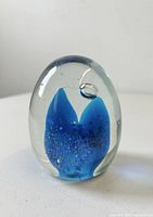 Clear ovoid glass paperweight featuring an internal blue abstract form and air bubbles, standing upright.