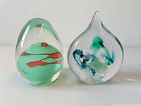 Front view of both paperweights showing egg and teardrop shapes with colorful internal glass patterns.