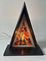 Front view of the triangular black wood lamp with colorful embedded glass fragments inside the pebbled lucite panel, lit to show glow.