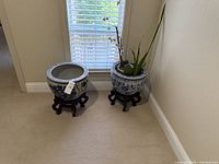 Two blue and white ceramic planters on carved wooden stands placed near window; one contains a plant