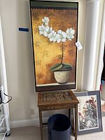 Wood carved Asian-inspired table with two framed floral paintings above and beside it, and a dark round container in front.