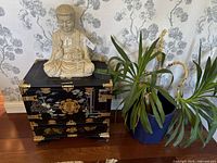 Front view of ceramic Buddha statue sitting on decorative wooden box, with green plant in blue pot adjacent