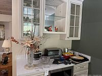 Wide view of white kitchen counter and cabinet holding various barware items and decor including a toaster oven, large crystal bowl, and tall crystal vase with dried flowers.