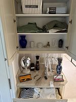 Open cabinet showing top shelves with bedding package, storage bag, folded textiles, decorative vases and canisters, countertop with decor and writing instruments
