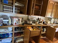 Wide view of kitchen cabinets and opened drawers showing linens, glassware, boxed kitchen appliance and cookware on counter and shelves