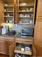 Kitchen cabinets open showing assorted dishware including glass, ceramic, cups, plates, and kettle on counter with Oster toaster oven and water dispenser.