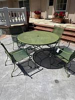 Full set of round green metal patio table with umbrella hole and four green metal mesh chairs on outdoor patio.