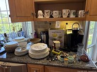 Overview of countertop showing disassembled kitchenware, dishware, ceramic mugs, a Keurig coffee maker, and water pitcher with various kitchen tools and small utensils laid out.