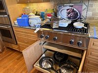 Full view of kitchen countertop and stove with assorted pots and pans, packaging, a blue cooler box, and cabinetry below open showing cookware.