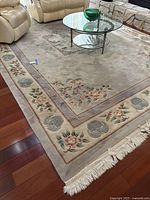Full corner view of the rug showing beige base color, bordered floral design, and fringed white edges on a wood floor next to beige leather recliner chairs and a glass coffee table.