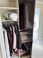 Full view of master bedroom hallway closet showing hung clothing, including dresses, coats, and fur coats on right side, with shelves holding folded bedding and bags.
