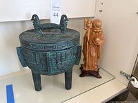 Full view of patina green metal ice bucket with intricate ancient Chinese style engravings, three legs, and lid with bird-shaped handle next to carved wooden statue.