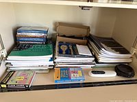Stack of piano and music books, flash cards, small clock, white remote, and a brown decorative item on a shelf.