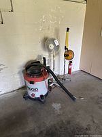 Photo of Ridgid shop vac, standing fan, floor lamp, and fire extinguisher against a wall on concrete floor.