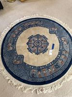 Full view of the round carpet showing beige center, blue border with motifs, and white fringes on edges.