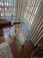 Two round glass top side tables placed near window blinds on wooden flooring. Tables have clear glass tops and metallic legs with circular support rings.