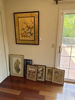 Wide view of seven framed art pieces arranged from large to small, including the large Asian botanical painting and smaller framed prints leaning against the wall near sliding glass door.