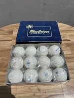 Open box showing 12 white MacGregor softballs individually wrapped in plastic, arranged in three rows of four.