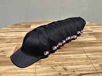 Photo showing 16 black mesh baseball caps lined up on wooden surface, each with an MLB hologram sticker on the brim
