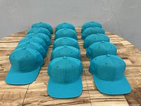 Group of 16 identical teal blue baseball caps arranged front-facing on a wooden surface.