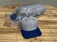 Sixteen gray and blue two tone baseball caps stacked and one in front for display.