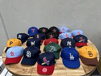 Front view of 20 assorted baseball caps on wooden table showing various MLB team logos.