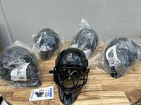 Five black Cooper 1000 catcher's helmets, four still in original plastic packaging, displayed on a wooden surface against a neutral wall.