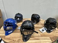 Group of 5 Cooper catcher's helmets on a wooden table showing different colors and styles.