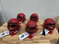 Group of 5 new red baseball catcher's helmets with metal face guards arranged on a wooden surface showing front and side views.