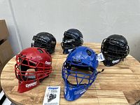 Group of 5 Cooper catcher's helmets in various colors displayed on wooden table, showing front and top views with tags attached.