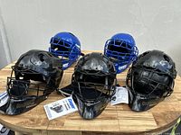 Five new catcher's helmets in black and blue with tags on wooden table.