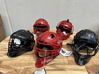 Group of five catcher’s helmets on wooden table, three red Cooper 1000 and two black Adams helmets, all new with tags.