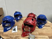 Group of five catcher's helmets showing three blue and two red helmets on a wooden table; two red helmets in front, with attached tags.