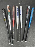 Full view of six aluminum baseball bats laid side-by-side on pavement showing all bats' handles and barrels.