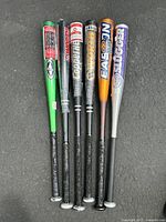 Six aluminum baseball bats laid side by side on black surface, showing full length including grips and barrels.