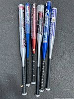 Group of six aluminum baseball bats with various brands and colors arranged on asphalt surface.