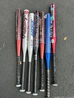 Photo showing six aluminum baseball bats in various brands lined up on concrete. Colors include metallic red, silver, blue, black and white with decals and grip handles.