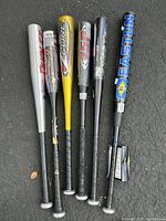 View of six aluminum baseball bats standing upright on pavement showing brands and colors