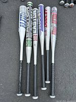 Six aluminum baseball bats laid out on the ground showing full length from grip to barrel, identifiable brands and models visible