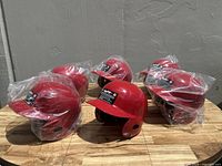 Photo showing six Rawlings red batting helmets on a wooden surface, mostly wrapped in plastic packaging.