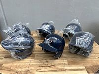 Group of six navy Rawlings batting helmets wrapped individually in plastic, arranged on wooden floor.
